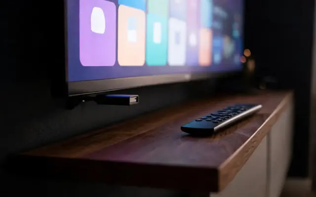 A streaming stick plugged into the side of a wall-mounted TV, its remote on the TV unit below